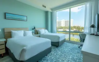 Hotel room with two beds, a large window boasting a city view, a desk, and a TV. The decor features light blue walls and a patterned carpet. Our commitment to institutional maintenance ensures the highest standards of cleanliness and comfort throughout your stay.