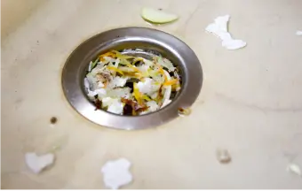 A kitchen sink drain filled with food scraps, including egg shells and vegetable peels, on a stained countertop.