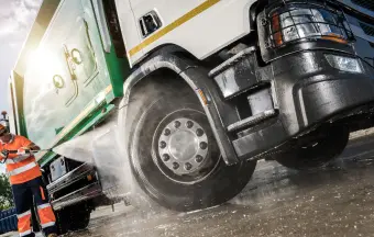 A sanitation worker standing next to a garbage truck being washed, focusing on the truck’s front wheel and splashing water.